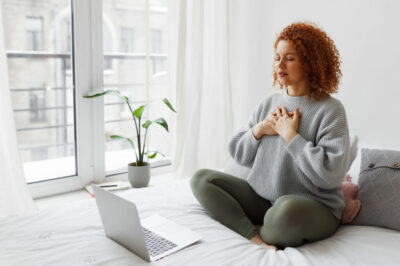 Pretty redhead plus size female sitting on bed in lotus posture with hands on chest doing pranayama techniques in front of laptop, breathing deeply, reaching inner balance after stressful day